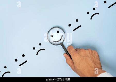 A hand holding a magnifying glass over a blue background with drawings of various facial expression. The magnifying glass focuses on one happy face. Stock Photo