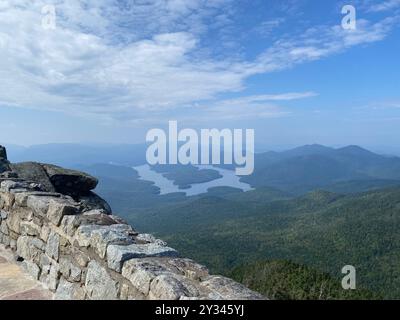 A scenic view from the summit of Whiteface Mountain in New York, showing a stone wall and a winding river amidst lush green forest under a blue sky. Stock Photo