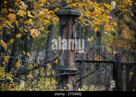 An old rusted metal gate post entwined with barbed wire, surrounded by autumn foliage, evoking a sense of abandonment. Stock Photo