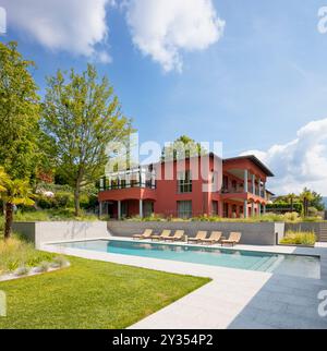 Red-walled villa in a big green garden with trees. There is a big swimming pool and sun loungers. Sunny summer day. Nobody inside Stock Photo