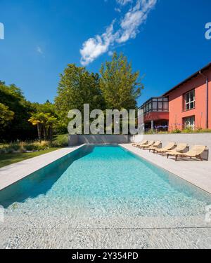 Red-walled villa in a big green garden with trees. There is a big swimming pool and sun loungers. Frontal view. Sunny summer day. Stock Photo