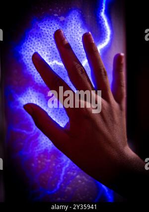 Close-up of a hand touching a glowing blue plasma ball, creating an electric light effect. Stock Photo