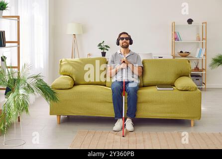 Portrait of blind man sitting on sofa at home in headphones and listening to music or audiobook. Stock Photo