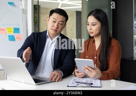 Asian business professionals engaged in discussion using laptop and tablet in office. Teamwork and collaboration for project planning. Display of focus, strategy, and communication skills Stock Photo