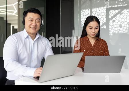 Asian business professionals using laptops and headsets working together in modern office setting. Collaborative teamwork, productivity, and communication emphasized. Stock Photo