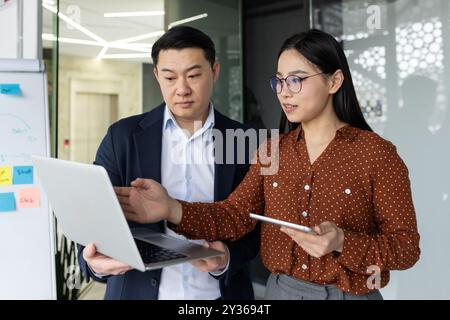 Asian business team working together in office environment. Man and woman discussing project using laptop and holding tablet. Professional interaction showcases teamwork, technology integration Stock Photo