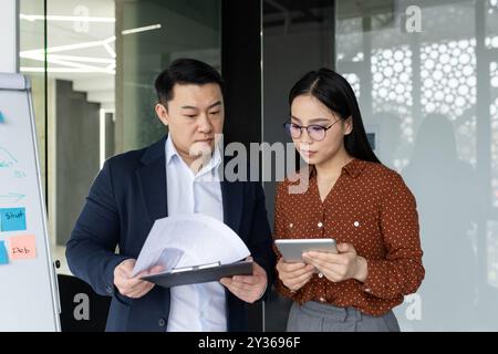 Asian business team focused on collaborative work in office setting. Man holds documents, woman uses tablet for analysis. Engaging in strategic planning and data examination. Concepts of teamwork, technology, and communication. Stock Photo