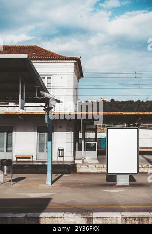 A vertical shot of a blank mockup billboard on a deserted railway platform in a suburban area. Old station building with metal roof, blue pillar, and Stock Photo