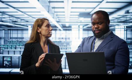 Teamworking technicians managing data center with advanced technology using devices. Coworkers in server room optimizing data center infrastructure with laptop and tablet, camera B Stock Photo