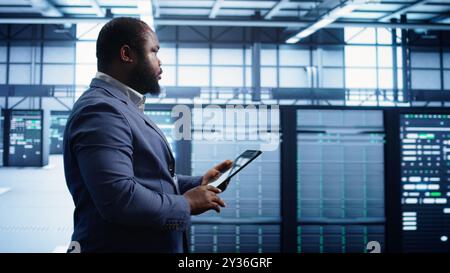 Technician walking in server room, doing checkup, inputting data on tablet of needed fixes. IT professional working in data center facility housing storage infrastructure, camera B Stock Photo