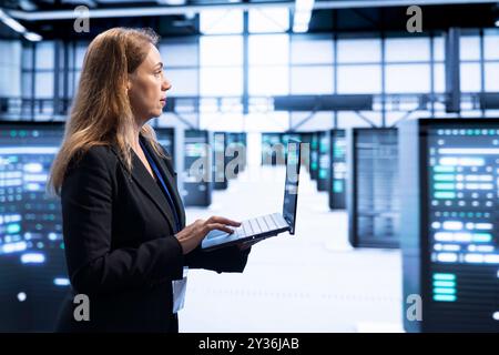 IT staff member walking in data center, using laptop to configure network infrastructure such as routers, switches and firewalls. Woman working in server room looking for technical issues Stock Photo
