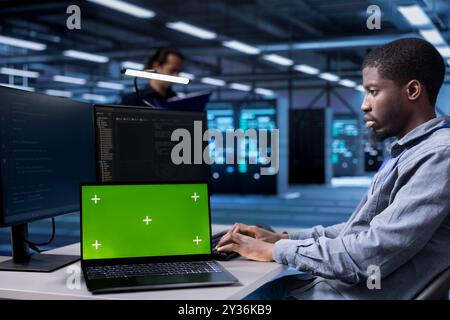 Engineer in data center running system diagnostic tests using isolated screen laptop. African american man in server farm deploying error checking utilities on mockup notebook to repair network issues Stock Photo
