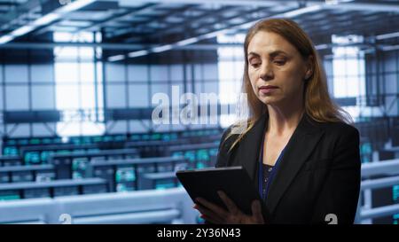 Admin in server room checks recovery plan on tablet, monitoring equipment energy consumption. IT professional uses notebook in data center workplace, ensuring rigs are functioning optimally, camera A Stock Photo