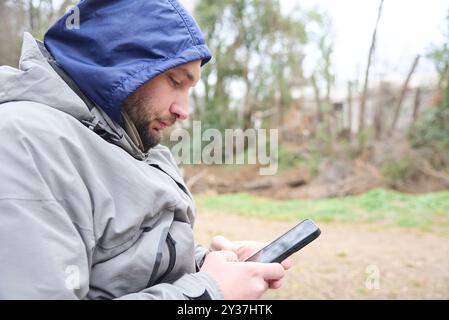 Side portrait of a young man dressed in warm clothes using his cell phone outdoors. Composition with copy space. Stock Photo