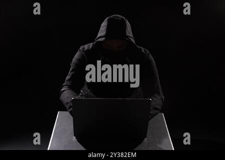 Cyber hacker in hood behind laptop in dark room, digital cyber security concept, computer hacking, information security Stock Photo