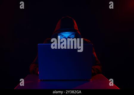 Cyber hacker in hood behind laptop on dark background with red blue light, anonymous spy. digital cyber security concept Stock Photo
