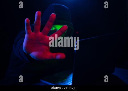 Cyber hacker in hood behind laptop puts hand forward hiding his face in dark room, concept of digital information protection, Computer hacking, stop c Stock Photo
