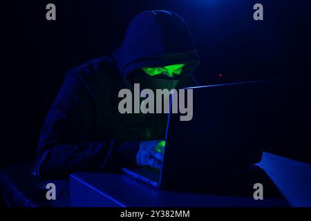 Cyber hacker in hood behind laptop in dark room, digital cyber security concept, computer hacking, information security Stock Photo