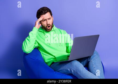 Photo of handsome young guy sit beanbag hold netbook nervous worried dressed stylish green garment isolated on violet color background Stock Photo