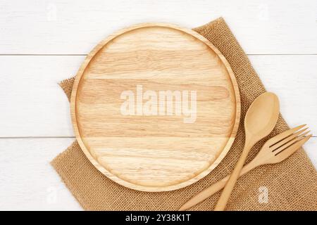 Empty round wooden plate with spoon, fork and rustic brown burlap cloth on white wooden table. Top view image. Stock Photo