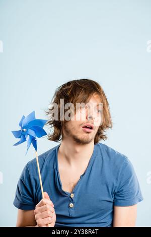 Bored, pinwheel and man in studio with toy isolated on blue background space. Energy, windmill and childish person play with plastic origami fan for Stock Photo