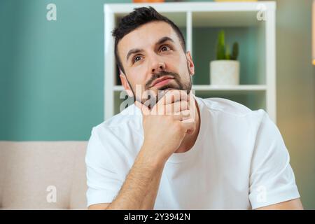 Thoughtful young man facing the camera while leaning forward on the sofa  Stock Photo