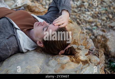 woman is lying eyes closed on a rocky surface with hand on forehead Stock Photo