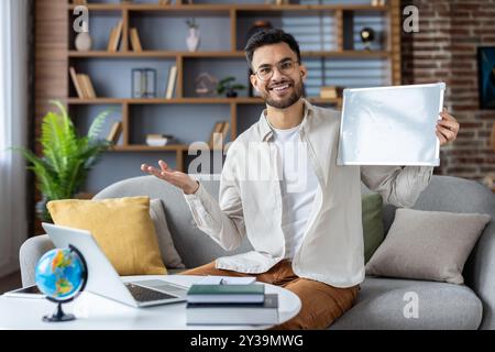 Smiling teacher holding blank whiteboard in home office. Engaged in online lesson with laptop and globe on desk. Cozy and inviting environment for teaching and learning Stock Photo