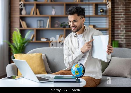 Smiling man engaged in online teaching session holds blank whiteboard while using laptop. Concept of remote education, virtual communication, and modern learning in home office environment. Stock Photo