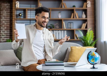 Cheerful online teacher conducting virtual class from home. Man with glasses gesturing while holding notebook, engaging with students through laptop. Stock Photo