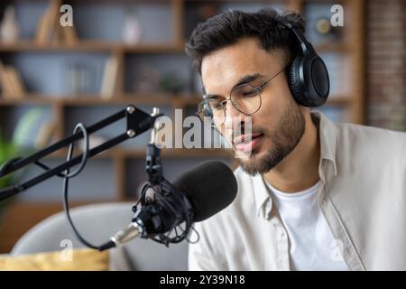 Young man wearing headphones speaking into microphone in podcast studio. He appears focused and engaged in serious conversation. Modern equipment and professional setup suggest tech-savvy environment Stock Photo
