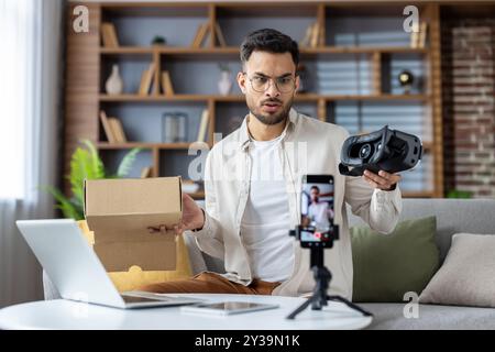Man exploring virtual reality headset usage in home workspace setting. Capturing video content with smartphone for online review. Engaging in modern technology trends and digital content creation. Stock Photo