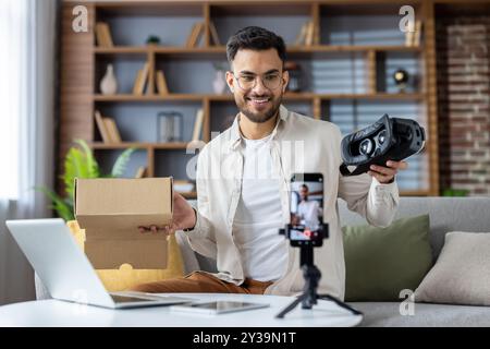 Young man creating unboxing video using smartphone. He holds virtual reality headset and cardboard box, capturing engaging content in home setting. Enthusiastic expression shows excitement about tech Stock Photo