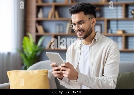 Young man with glasses using tablet in cozy living room. Content creator or student happily browsing digital content on device. Relaxed atmosphere enhanced contemporary decor and natural lighting. Stock Photo