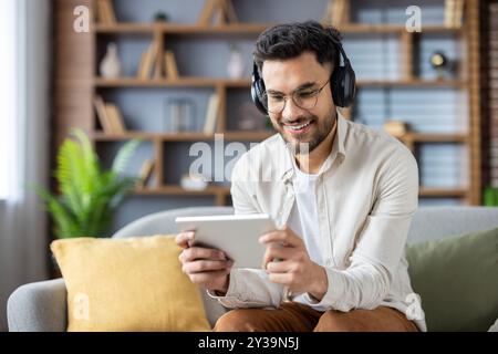 Cheerful man wearing headphones and casual clothes sits on sofa using digital tablet for entertainment or education. Relaxed, happy atmosphere with modern home setting enhances tech-focused lifestyle Stock Photo