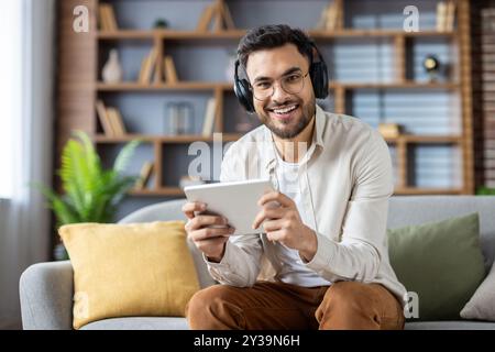 Hispanic man sitting comfortably on couch using tablet and headphones. Smiling, he enjoys digital entertainment at home. Relaxed atmosphere indicates a peaceful and fulfilling moment of leisure Stock Photo