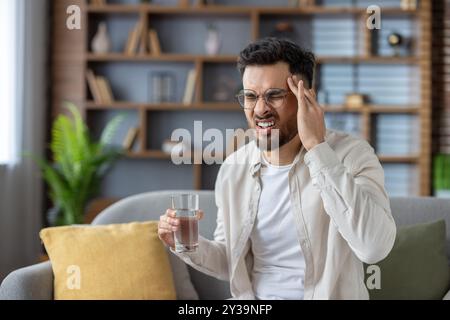 Young man experiencing headache sitting on sofa indoors holding glass of water. Person showing discomfort and pain, possibly from stress or fatigue, seeking relief and comfort in home environment. Stock Photo