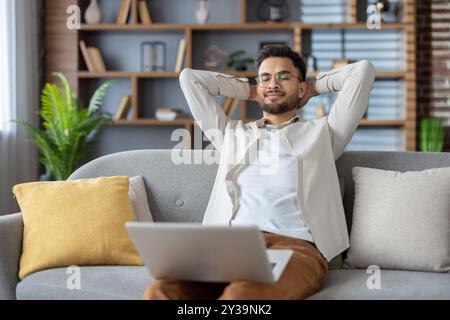 Young relaxed Indian man sitting on sofa at home, holding laptop on lap, arms behind head and closing eyes resting. Stock Photo