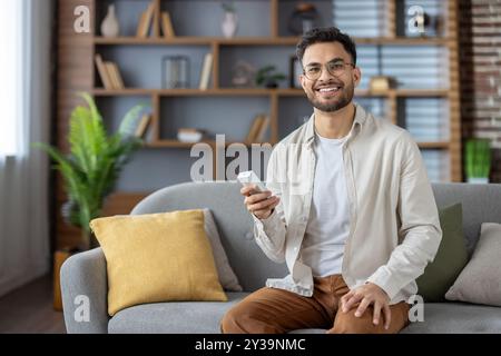 Smiling hispanic man sitting on couch holds air conditioner remote control in hands. Relaxing at home, enjoying comfort and modern technology in cozy living room setting. Stock Photo