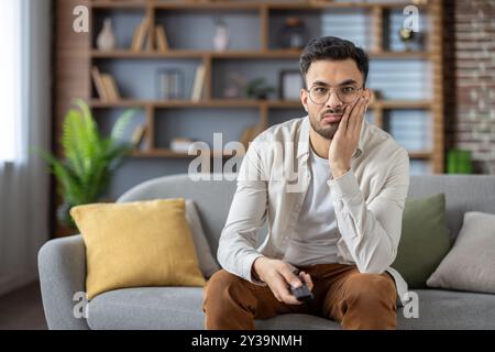 Tired and bored young indian man sitting in front of camera at home, holding head in hand and holding tv remote control. Stock Photo