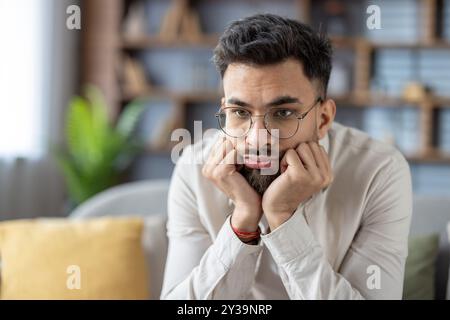 Young man wearing glasses sitting in living room appearing bored and thoughtful. Casual attire highlights relaxed atmosphere. Expression conveys mix of boredom and contemplation Stock Photo