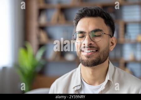 Young man with glasses smiling peacefully while meditating indoors. Capturing moment of relaxation, serenity, and mindfulness. Closed eyes convey sense of calm and inner peace tranquil environment. Stock Photo
