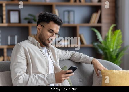 Man sitting comfortably on sofa using smartphone, casually dressed in light shirt. Relaxed atmosphere as he browses internet, checks emails, or engages on social media. Modern living room setting Stock Photo