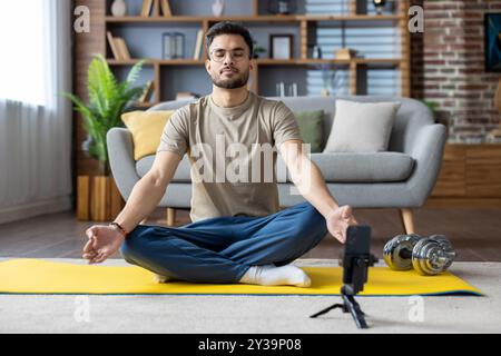 Indian young man doing online yoga at home, sitting on mat in lotus position with closed eyes in front of mobile phone. Stock Photo