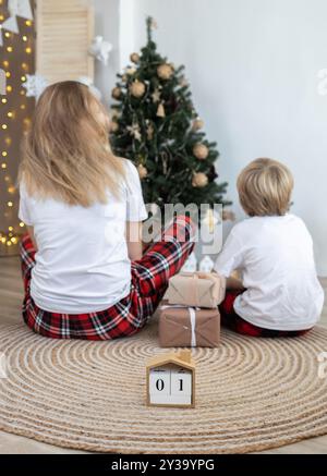 Mom and son sit with their backs on the floor in front of the New Year tree next to gift boxes. Festive atmosphere. Waiting for a Christmas miracle. F Stock Photo