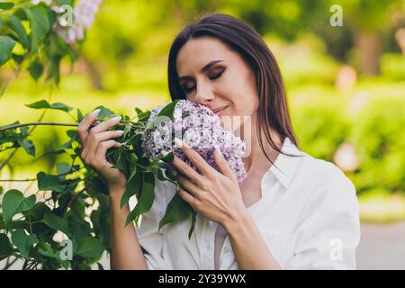 Photo of young cheerful woman posing smell lilac flowers closed eyes wear white outfit walk park sunny summer weather outside Stock Photo