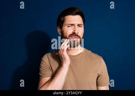 Photo of suffering man with stylish beard dressed beige t-shirt hold hand on cheek have toothache isolated on dark blue color background Stock Photo