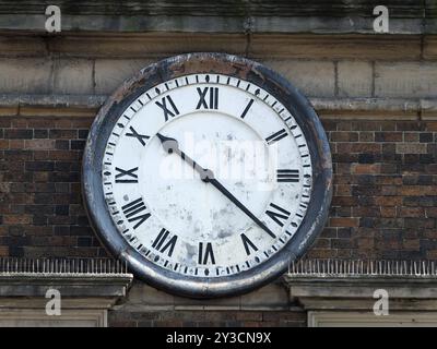 Old round clock on the brick wall of a building with roman numerals and peeling distressed face with hands at twenty past ten Stock Photo