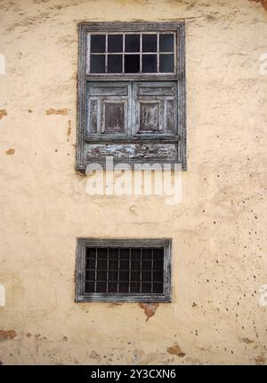 Two old wooden windows with ancient faded peeling green paint metal bars shutters and panels on an old plastered peeling concrete terracotta and beige Stock Photo