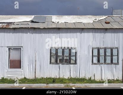 Full frame close up of a shabby old dilapidated corrugated iron building with with closed painted over patched windows repaired rusting roof and white Stock Photo
